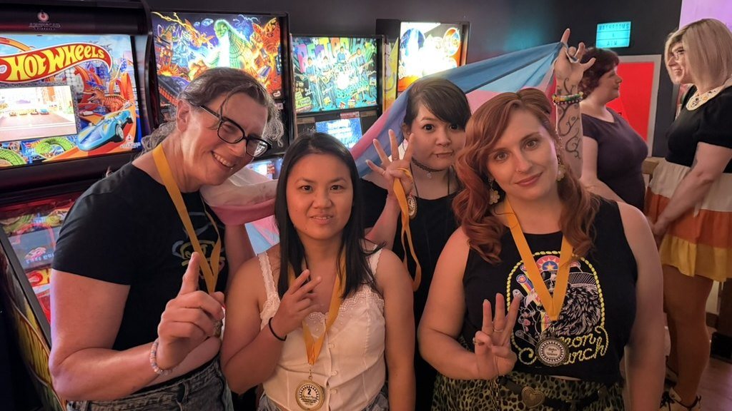Three women wearing gold medals hold up fingers for their placings in front of pinball machines, with a trans pride flag behind them